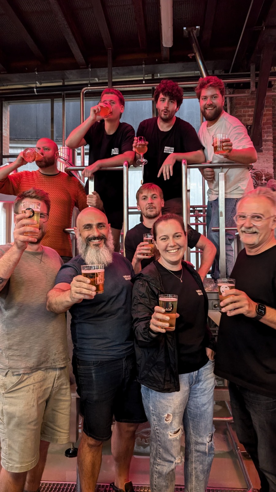 Foto colorida em espaço interno de cervejaria mostra nove pessoas, sendo oito homens e uma mulher, sorindo e brindando com copos de cerveja. Fim da descrição.